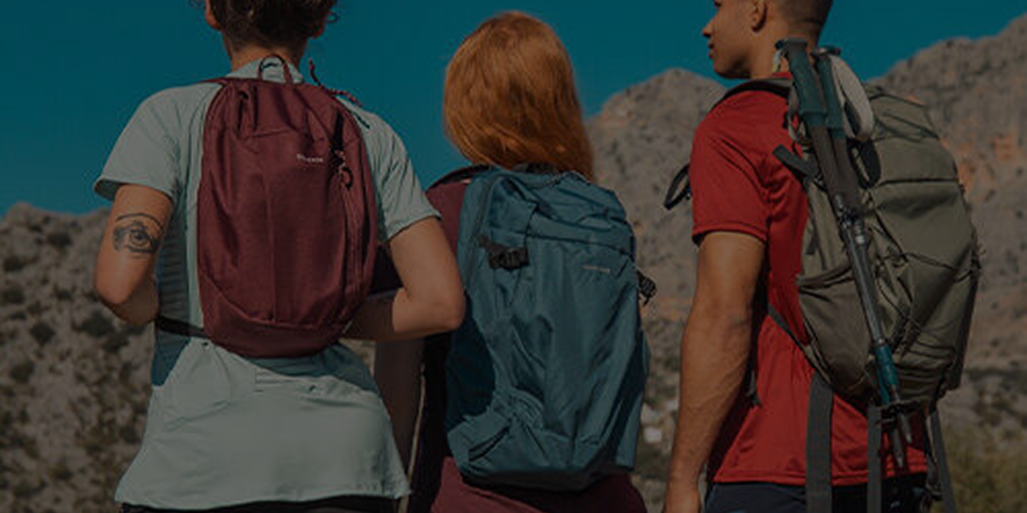 Equipo de montaña: tres mochilas técnicas en colores morado, azul y verde, y bastones de trekking en paisaje rocoso